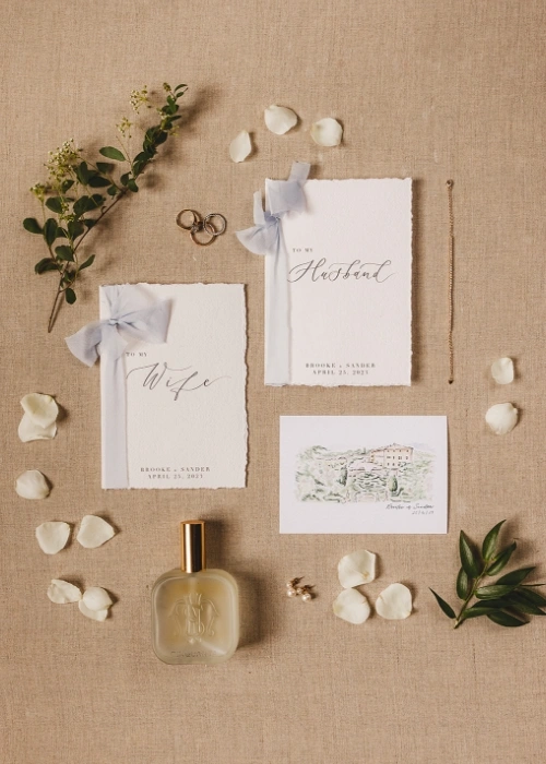 Detail shoot of vows booklet with venue drawing, wedding bands, perfume and flower petals