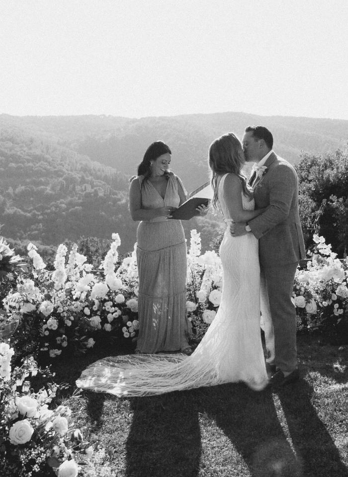 Bride and groom kiss during wedding ceremony surrounded by flowers and views of Tuscan hills