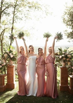 Bride and bridesmaids joyfully lift their bouquets at pink wedding ceremny in Tuscany