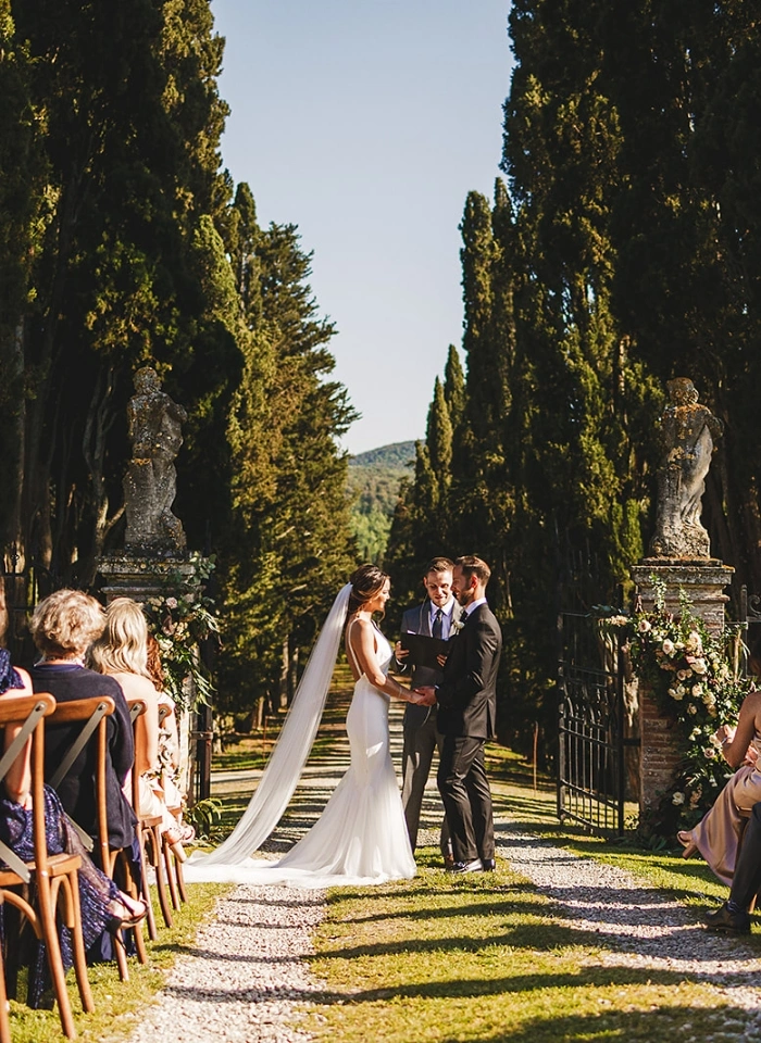 Bride and groom hold hands during outdoor wedding ceremony in front of Borgo Stomennano's gate