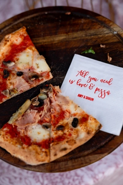 Pizza slices served on a table next to a personalized wedding napkin that reads "All you need is love & pizza."
